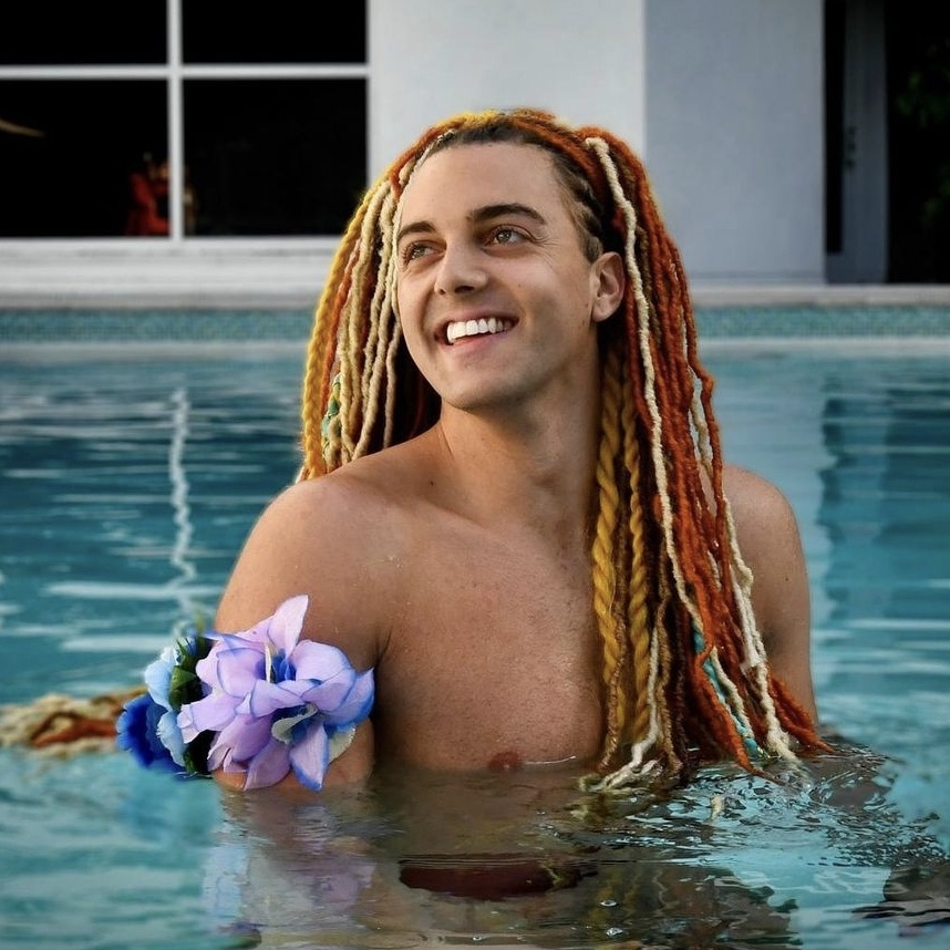 Prince Pōhaku smiling in a sunlit pool, bright hair and flowers beside him